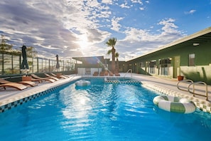 A swimming pool with lounge chairs and a sunshade, surrounded by a building with a green exterior. - Sagewater Springs - Adult Only (Desert Hot Springs)