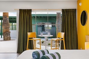 A hotel room with a bed, two chairs, a small table, and a view of a green building through large windows. - Sagewater Springs - Adult Only (Desert Hot Springs)