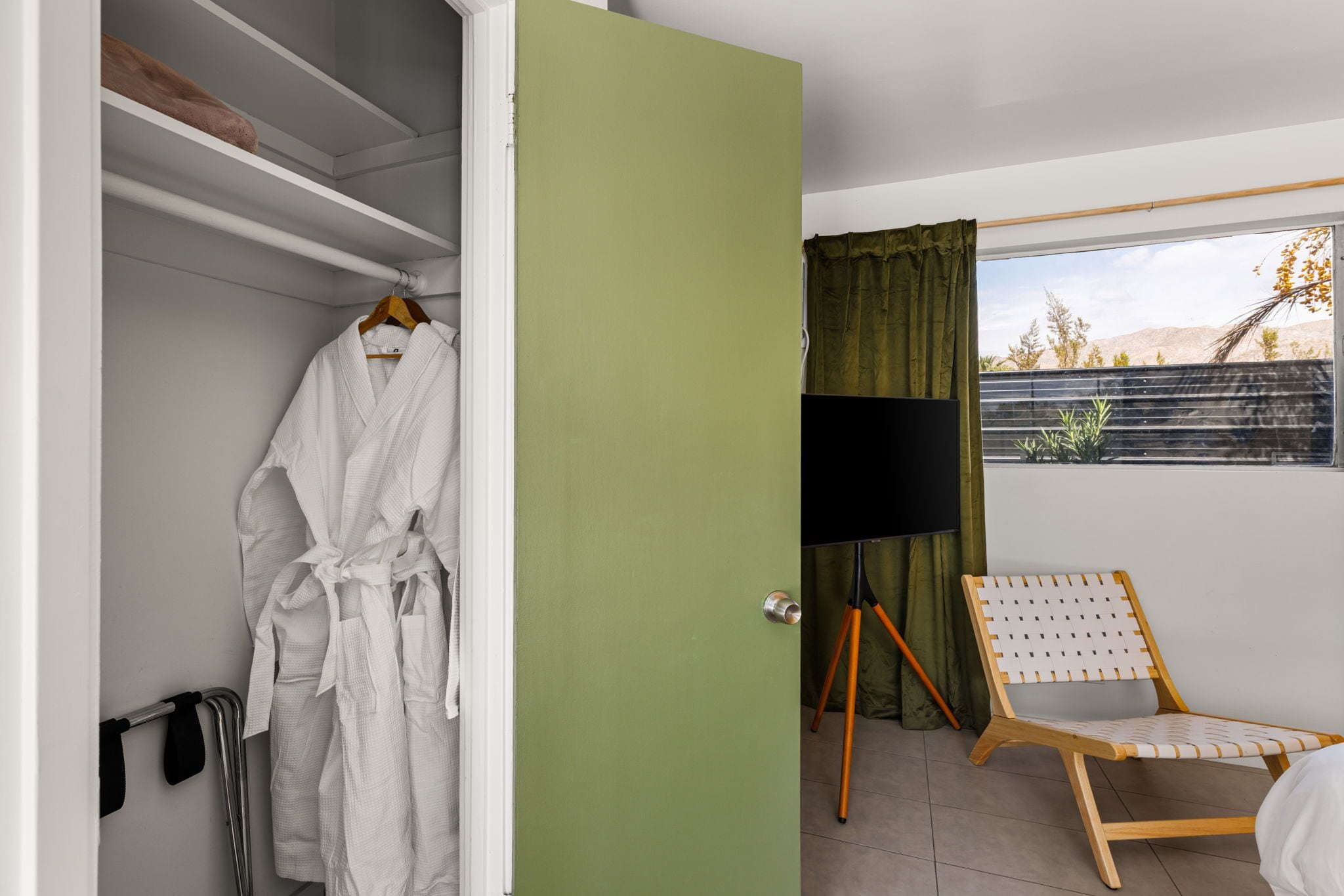 A hotel room with a walk-in closet, a television on a tripod, a chair, and a view of the outdoors.