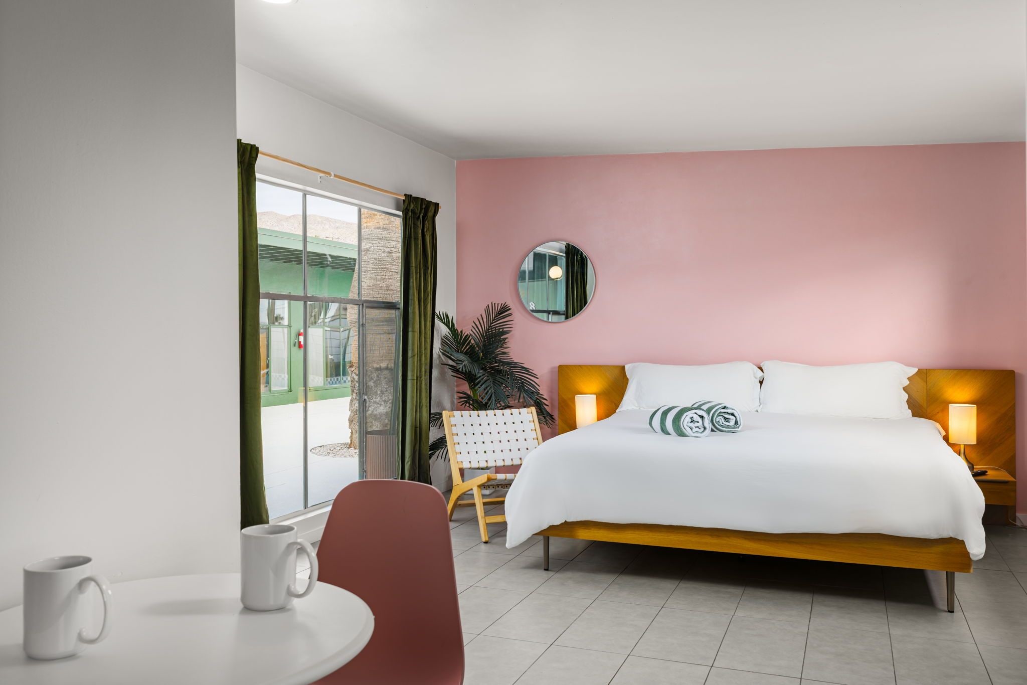 A hotel room with a bed, bedside lamps, a round mirror, a chair, a table with a mug, and a window with curtains.