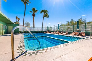 Una piscina rectangular con borde de baldosas a cuadros, rodeada de sillas de descanso y sombrillas, con palmeras al fondo.