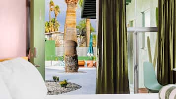 A hotel room with a bed, a view of an outdoor area with palm trees, a pool, and a green wall.