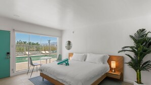 A bedroom with a bed, a bedside table with a lamp, a chair, a large window with a view of a pool and mountains, and a potted plant. - Sagewater Springs - Adult Only (Desert Hot Springs)