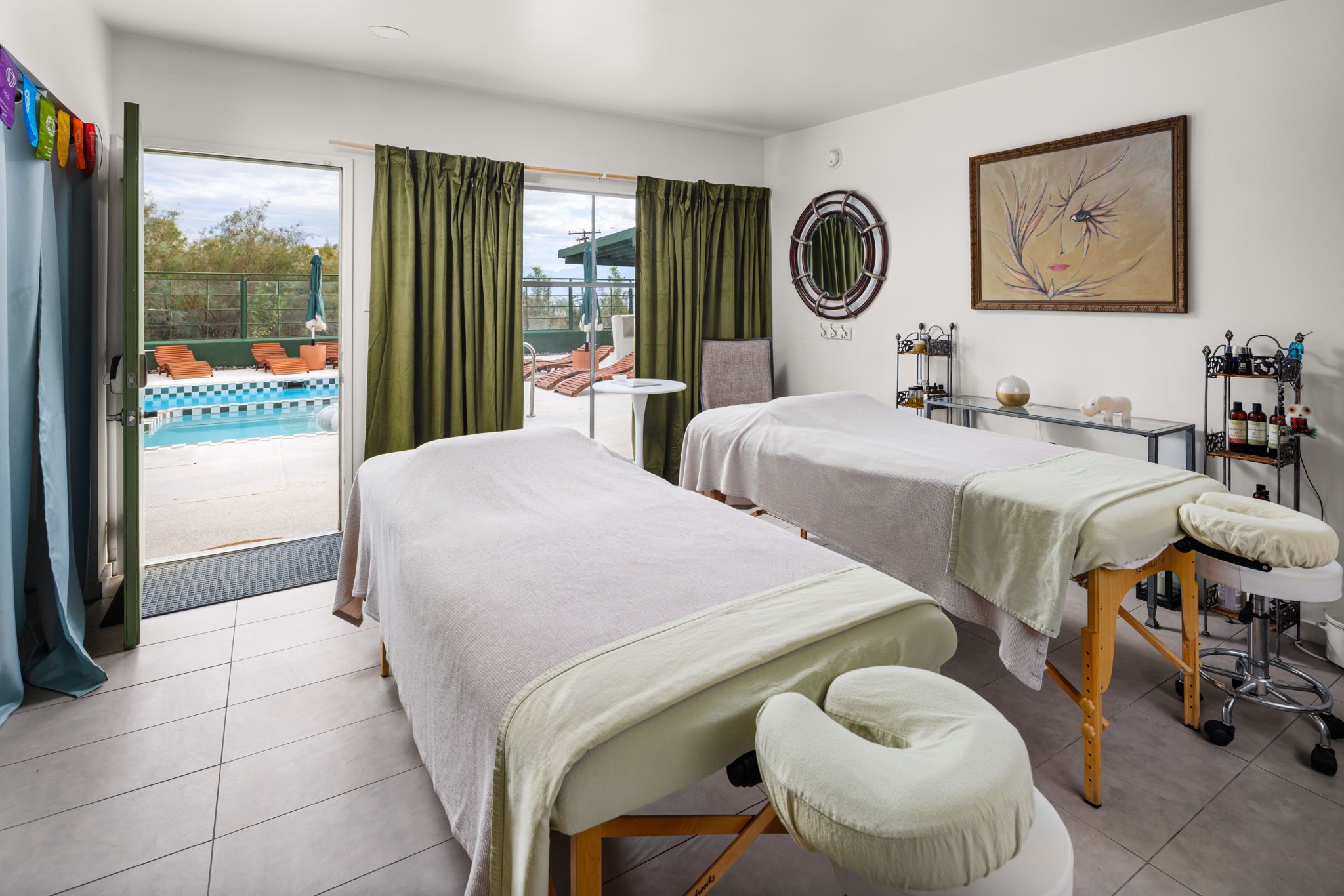 Une chambre d’hôtel avec deux lits, une table de massage, une petite table, une chaise, un tableau et une vue sur une piscine extérieure.
