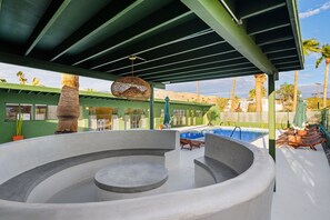 Un jacuzzi circular con piscina aledaña, un área de estar techada y vista a palmeras y montañas al fondo.