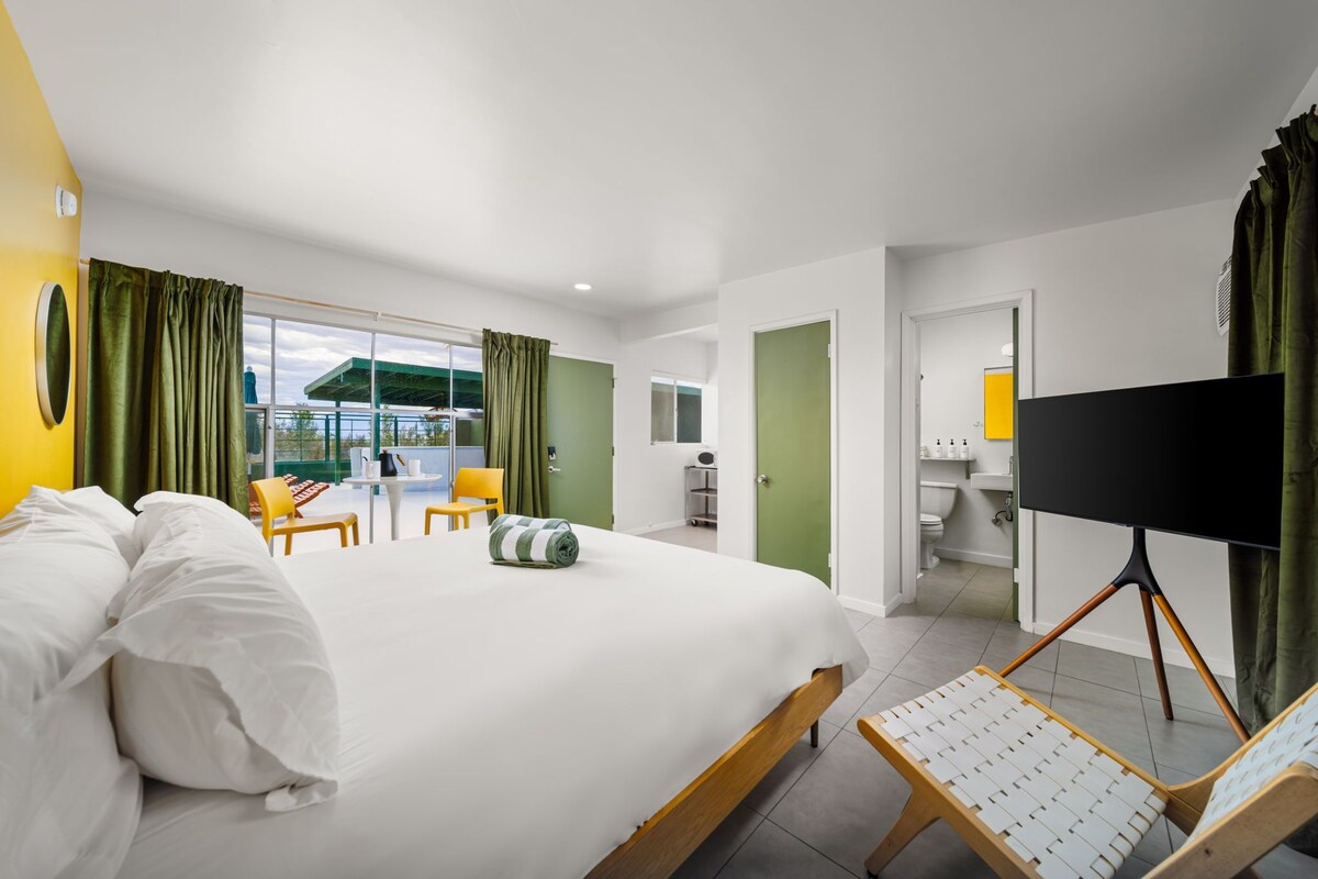 A hotel room with a large bed, a seating area with chairs, a TV on a stand, and a view of the outdoors.