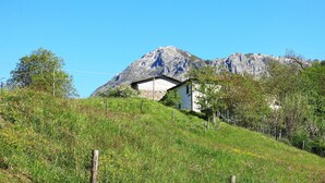 Une maison de campagne avec un toit en pente, entourée d’une colline verdoyante et adossée à une montagne.