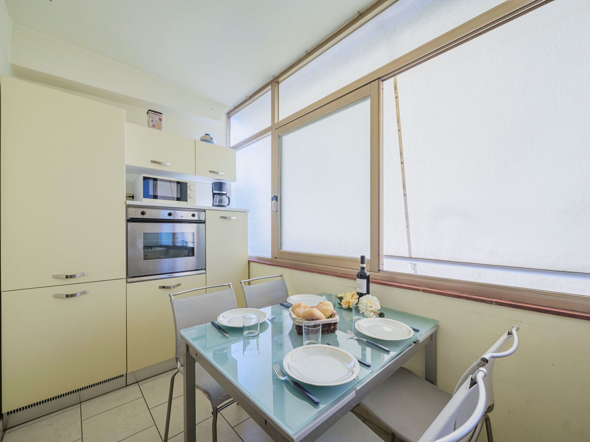 A compact kitchen with a microwave, oven, and coffee maker. There is a dining table with plates, glasses, and a basket of rolls.