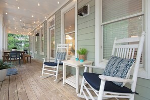 A covered porch with white rocking chairs and a table, featuring string lights and potted plants.