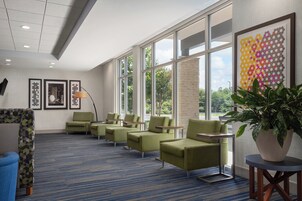 Un moderno vestíbulo de hotel con alfombra azul, sillones verdes y grandes ventanales con vistas al exterior.