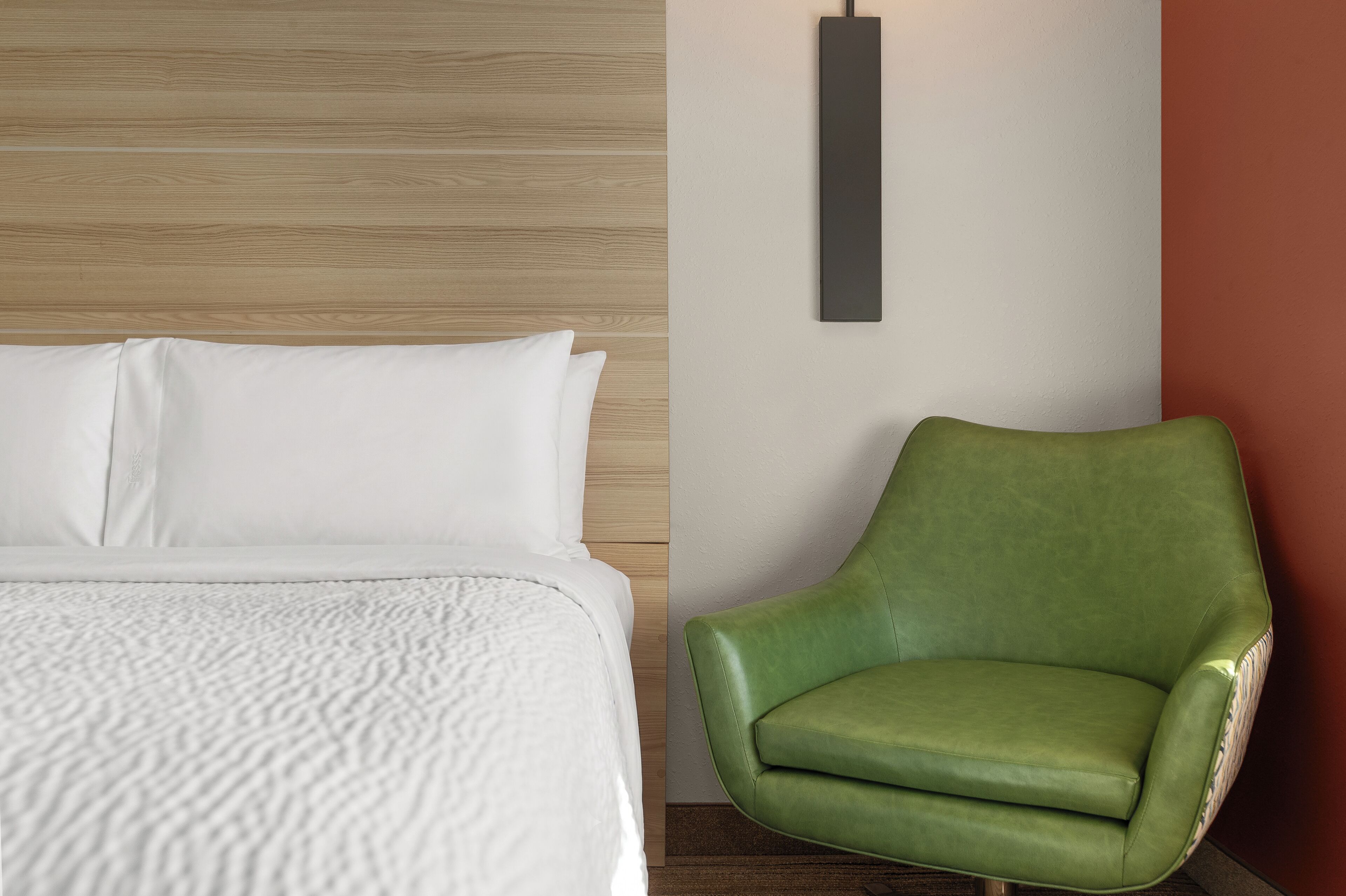 Una cama con ropa de cama blanca y una silla tapizada de verde frente a una pared con interruptor de luz.