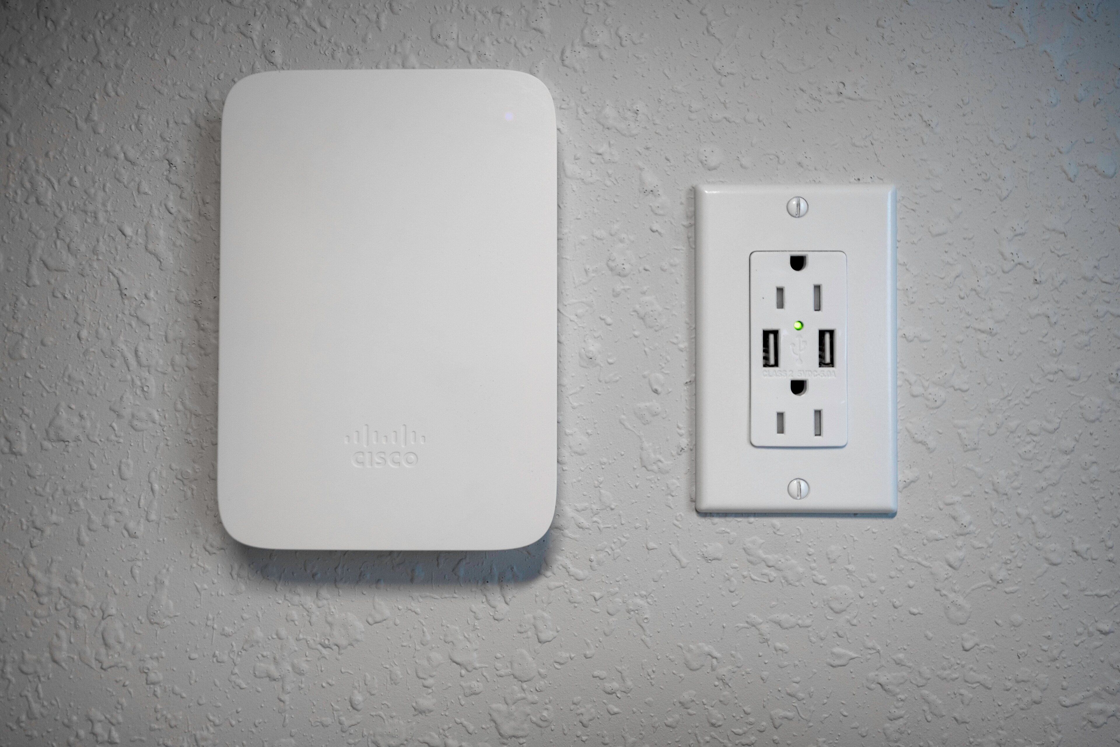 A white wall-mounted device with the logo 'Cisco' and a standard electrical outlet with two USB ports and a green indicator light.
