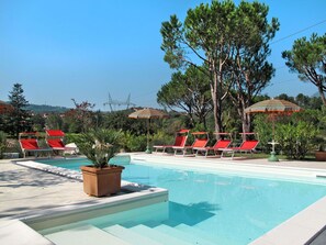 Ein Poolbereich mit roten Liegestühlen und gestreiften Sonnenschirmen, umgeben von Bäumen und blauem Himmel.