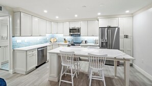 A modern kitchen with white cabinetry, a central island with bar stools, stainless steel appliances, and a blue-tiled backsplash.