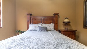 A bedroom with a wooden headboard, a bed with patterned bedding, a nightstand with a lamp, and a potted plant.