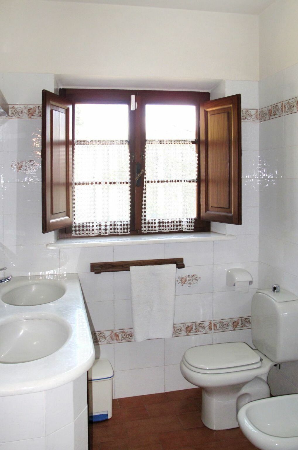 Une salle de bain avec deux lavabos, des toilettes et une fenêtre dont les volets sont ouverts.