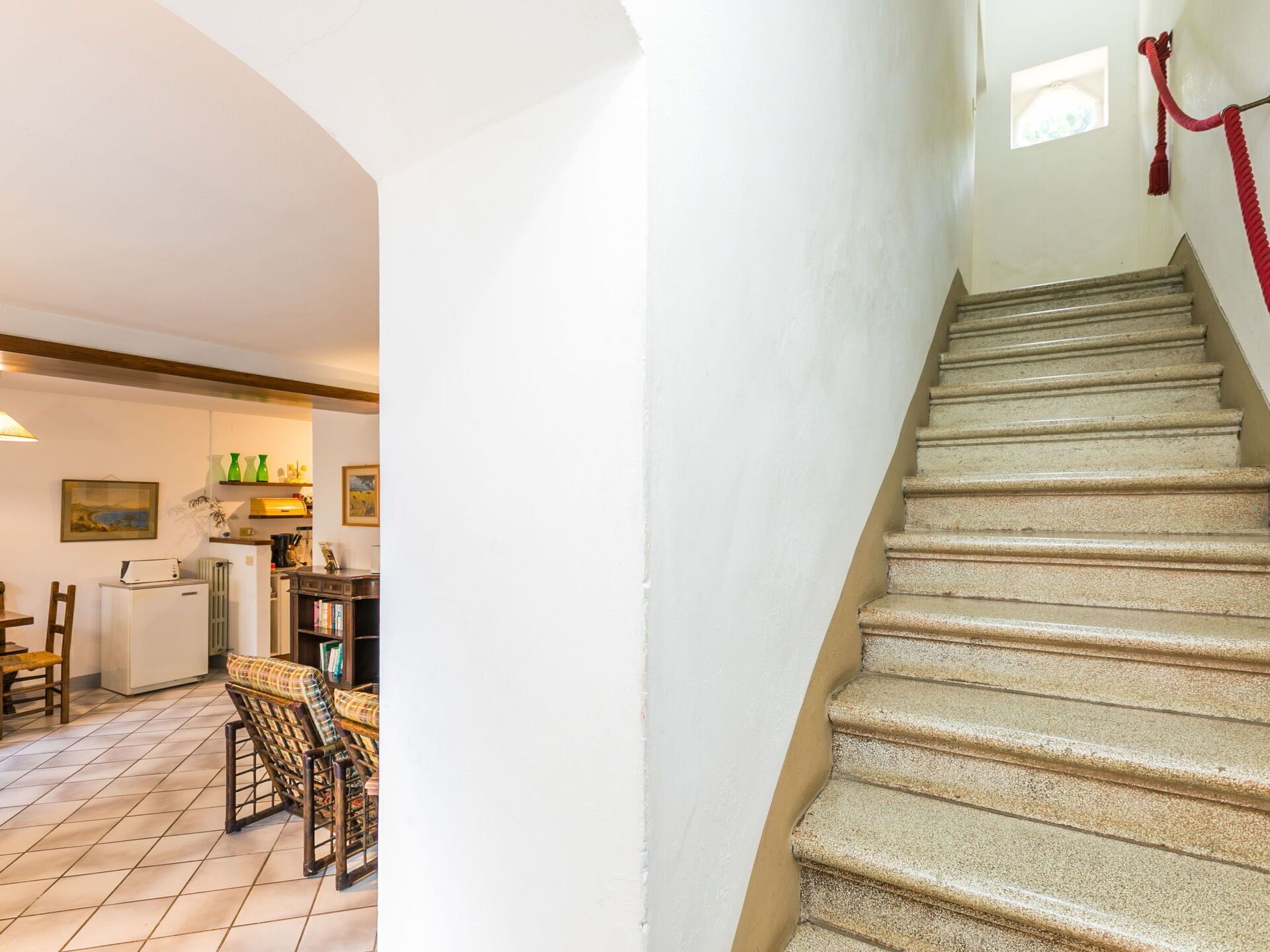 A staircase with a red rope barrier, a kitchen area with a refrigerator, a dining table with chairs, and a bookshelf.