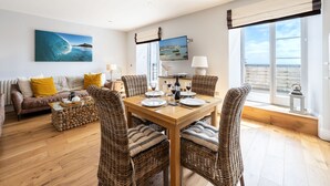 Un comedor con una mesa de madera dispuesta para cuatro personas, rodeado de sillas de mimbre, un sofá con cojines amarillos y un ventanal con vista al mar.