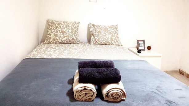 Uma cama bem arrumada com um cobre-leito estampado, duas almofadas e dois rolinhos de toalha em uma mesa de cabeceira com uma moldura e um pequeno objeto decorativo.