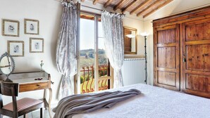 Un dormitorio con una cama grande, vigas de madera en el techo, un escritorio con una silla, un espejo y vistas a un viñedo a través de una ventana con cortinas.