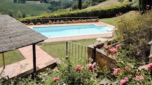 Una piscina con cubierta de madera y borde de piedra, rodeada por un jardín con flores rosadas y vistas a colinas ondulantes.