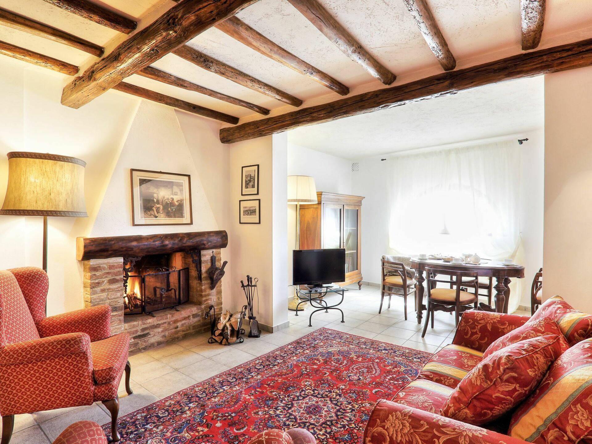 A cozy living room with a fireplace, red patterned sofas, a large rug, and wooden beams on the ceiling.