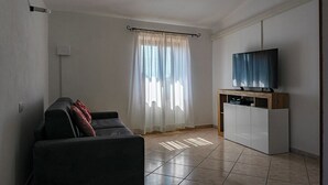 Ein Wohnzimmer mit einer Couch, einem Fernseher auf einem Holzschrank und einem Fenster mit durchscheinenden Vorhängen.