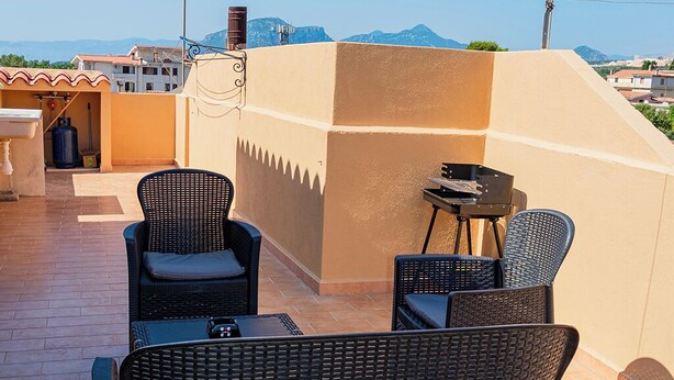Eine Terrasse im Freien mit Korbmöbeln und einem kleinen Tisch, mit Blick auf eine Stadtlandschaft und Berge im Hintergrund.