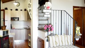 A cozy kitchen with a wooden stove, a white chair, and a dining table. There is a staircase with a metal railing leading to a landing with a bench and a decorative wall hanging. - Wonderful private villa for 2 people with WIFI, TV and terrace (Volterra)