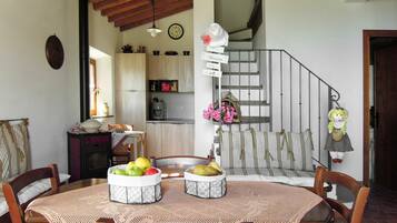 Un coin repas avec une table ronde, des chaises et un panier de fruits. On aperçoit une cuisine à l’arrière-plan, avec des placards et une cuisinière. Un escalier est visible sur la droite.