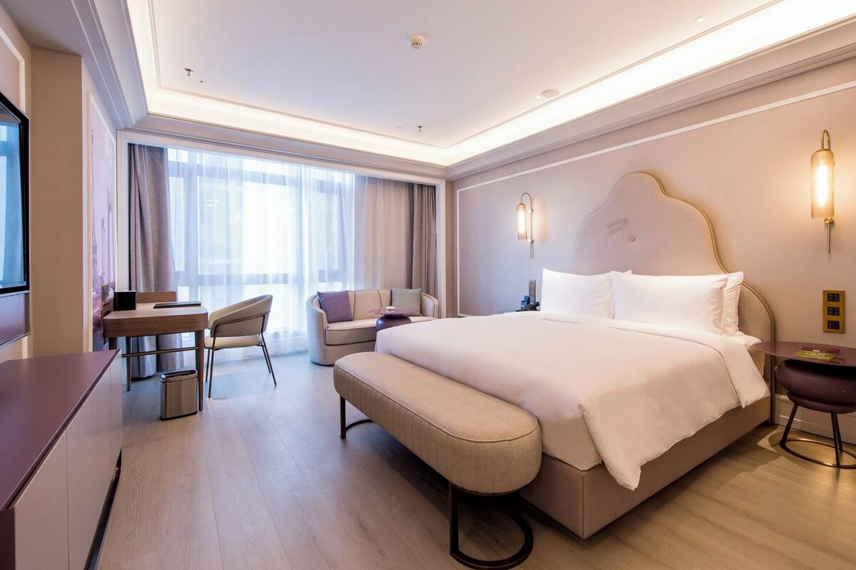 A hotel room with a large bed, a desk, a chair, a bench, and a small table.