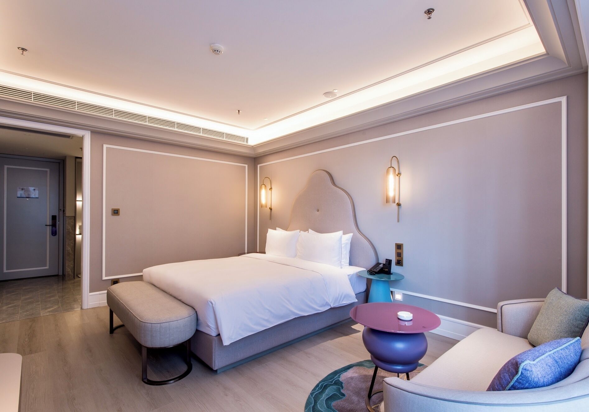 A modern hotel room with a large bed, a seating area with a round table, and a door leading to the hallway.