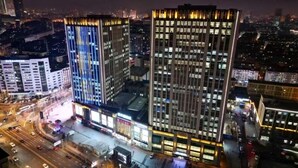 Un paisaje urbano nocturno con rascacielos, uno con fachada azul distintiva y otro con diseño decorativo.