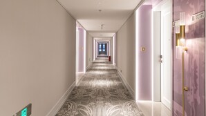 Un pasillo con alfombra estampada, una puerta con el número y un accesorio de luz en la pared.