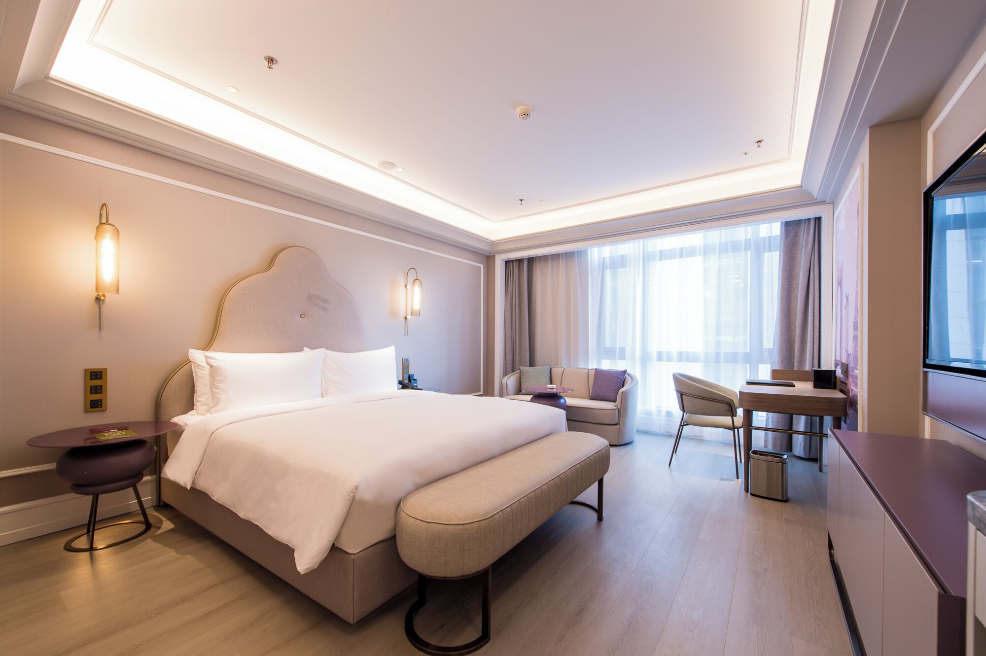 A modern hotel room with a large bed, a seating area with a sofa and armchairs, a small table, and a desk.