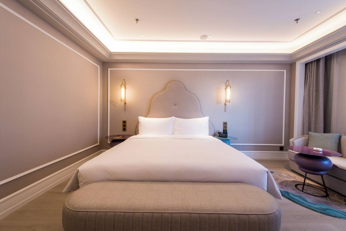 A large bed with white linens, two wall-mounted lamps, a nightstand, a small table, a chair, and a bench.