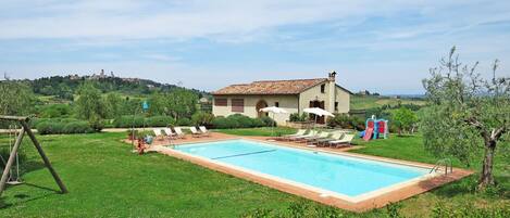 A swimming pool with lounge chairs, a small building with a tiled roof, and a grassy area with bushes and trees.