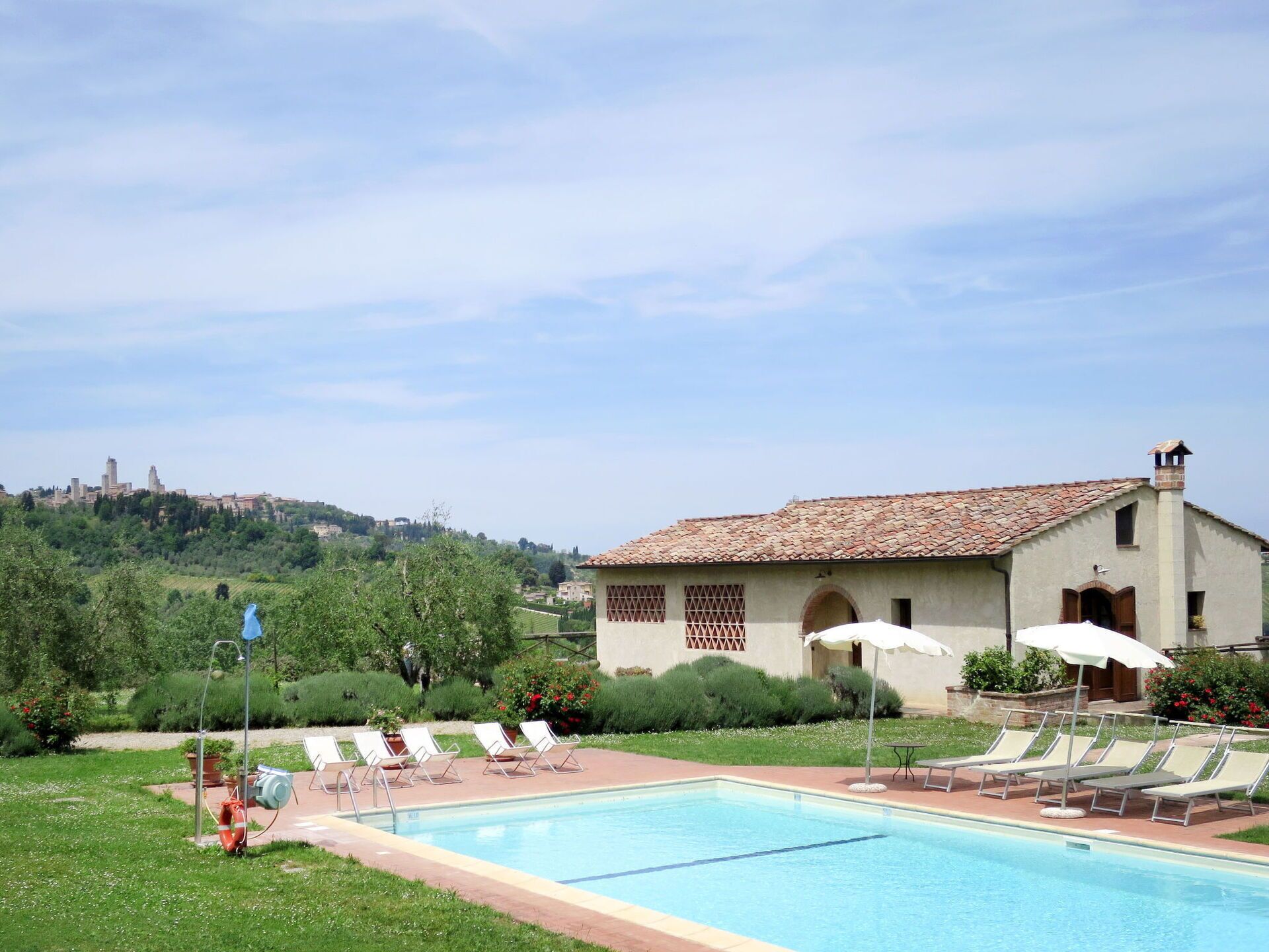 A swimming pool with lounge chairs and umbrellas, surrounded by greenery and a building with a tiled roof.