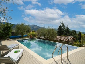 Ein Poolbereich mit klarem, blauem Wasser, einer Holzterrasse, Liegestühlen und Blick auf Berge im Hintergrund.