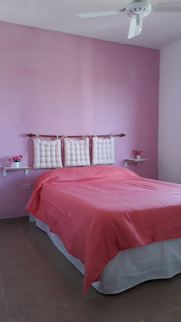 A bedroom with a pink wall, a bed with a red bedspread, two white pillows, a ceiling fan, and a small shelf with a vase of flowers.