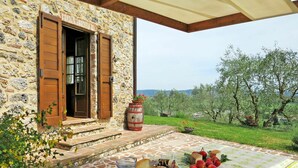 Un bâtiment en pierre avec une porte et des volets en bois, une table garnie de nourriture et de boissons, et une vue sur des arbres et des collines.
