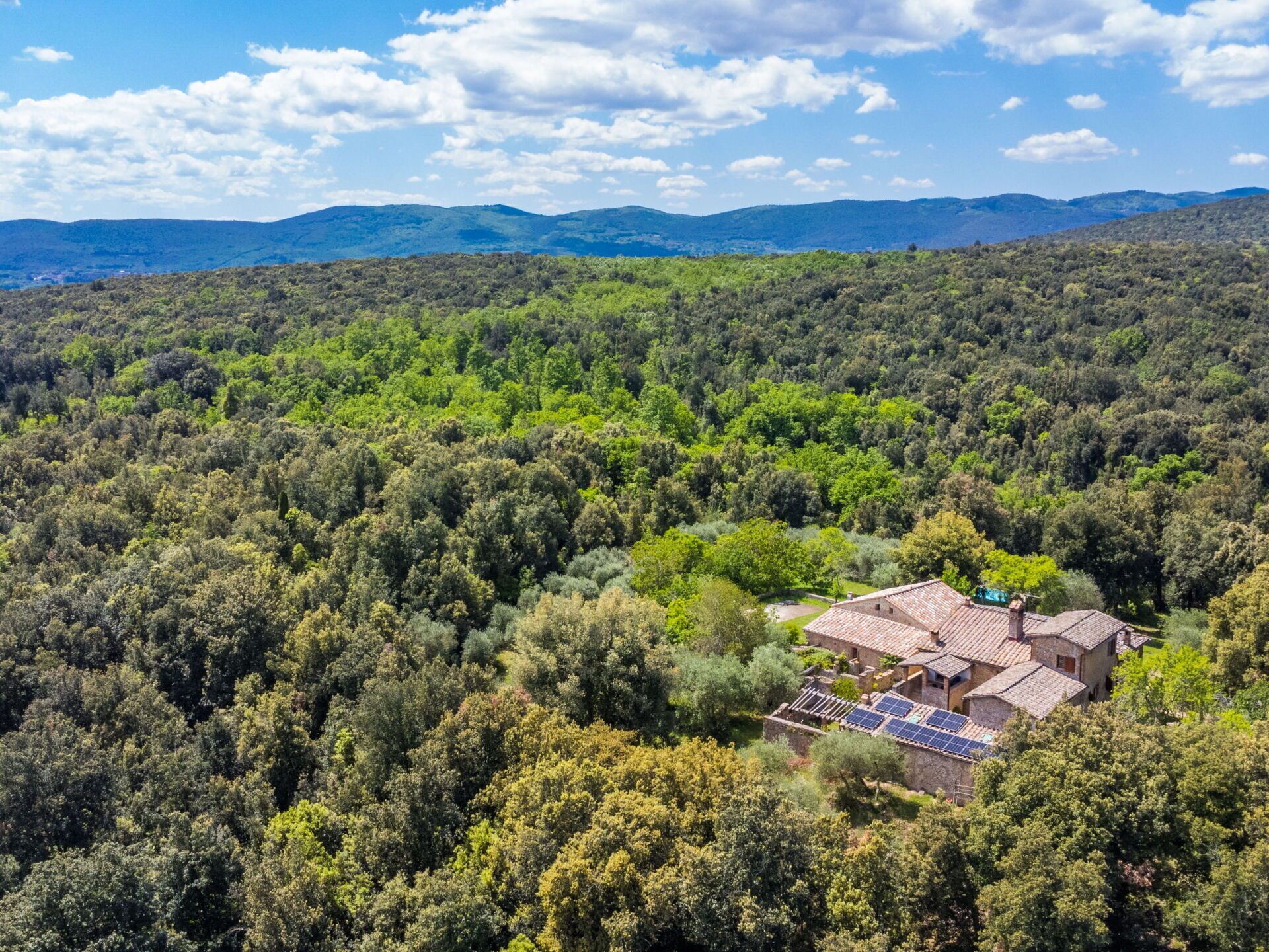 Une maison de campagne avec un toit en tuiles, entourée d’une forêt dense, dotée de panneaux solaires et offrant une vue dégagée sur les collines lointaines.