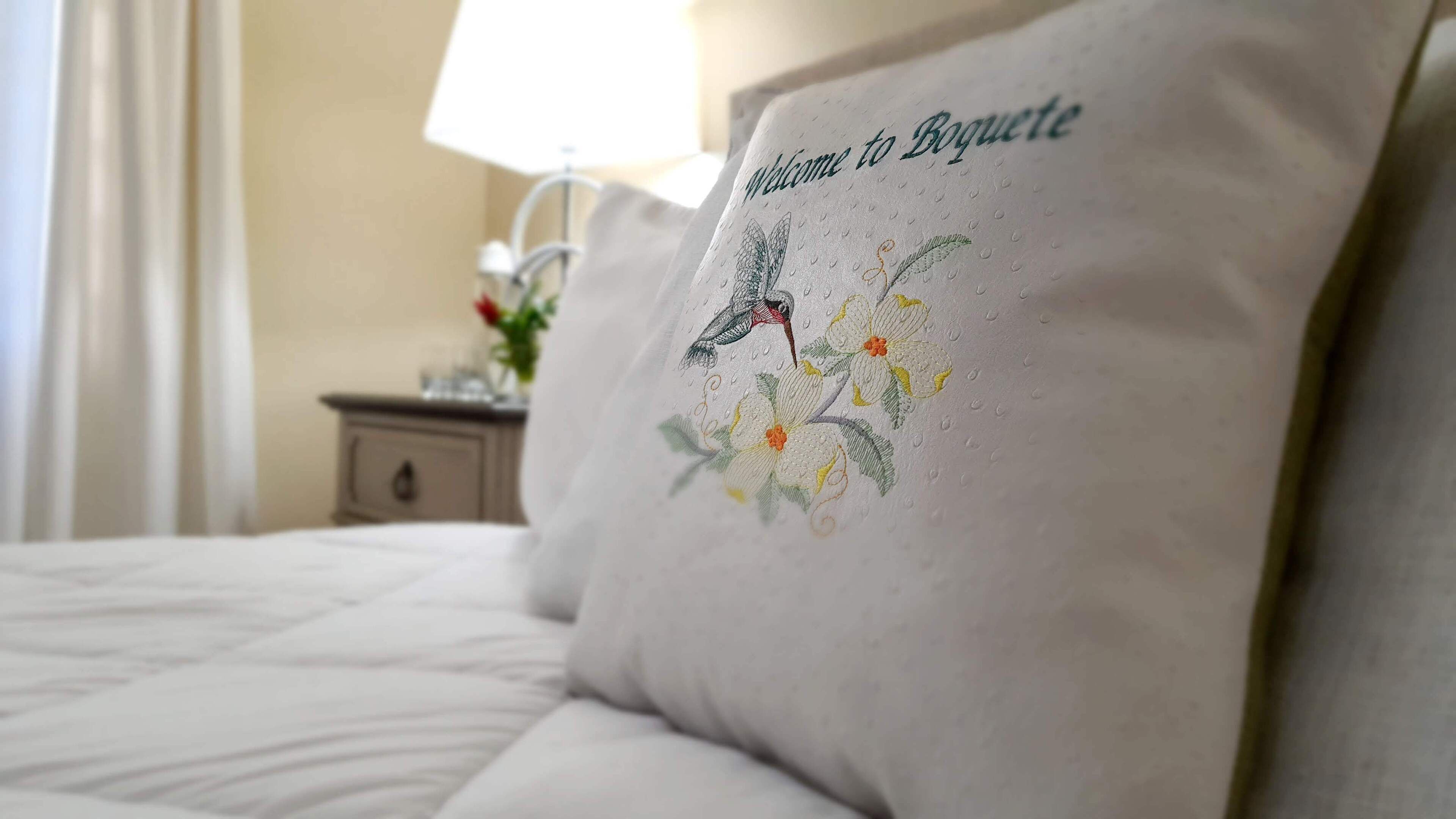 A hotel room with a neatly made bed, a bedside table with a lamp, and a pillow with a bird and floral design.