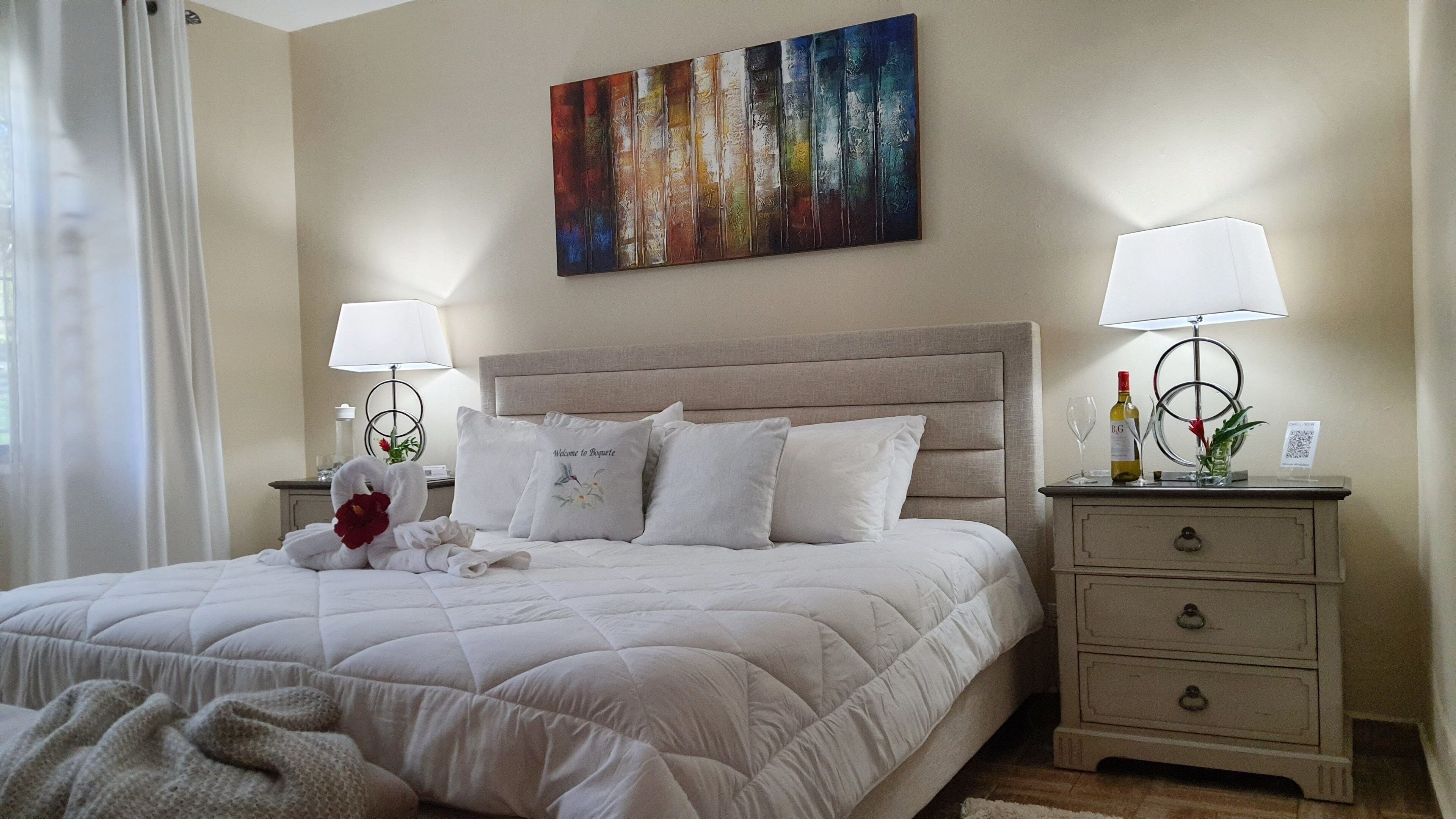 Una cama bien hecha con edredón blanco acolchado, dos mesitas de noche con lámparas, un cuadro decorativo en la pared y una mesita de noche con una botella y un vaso.