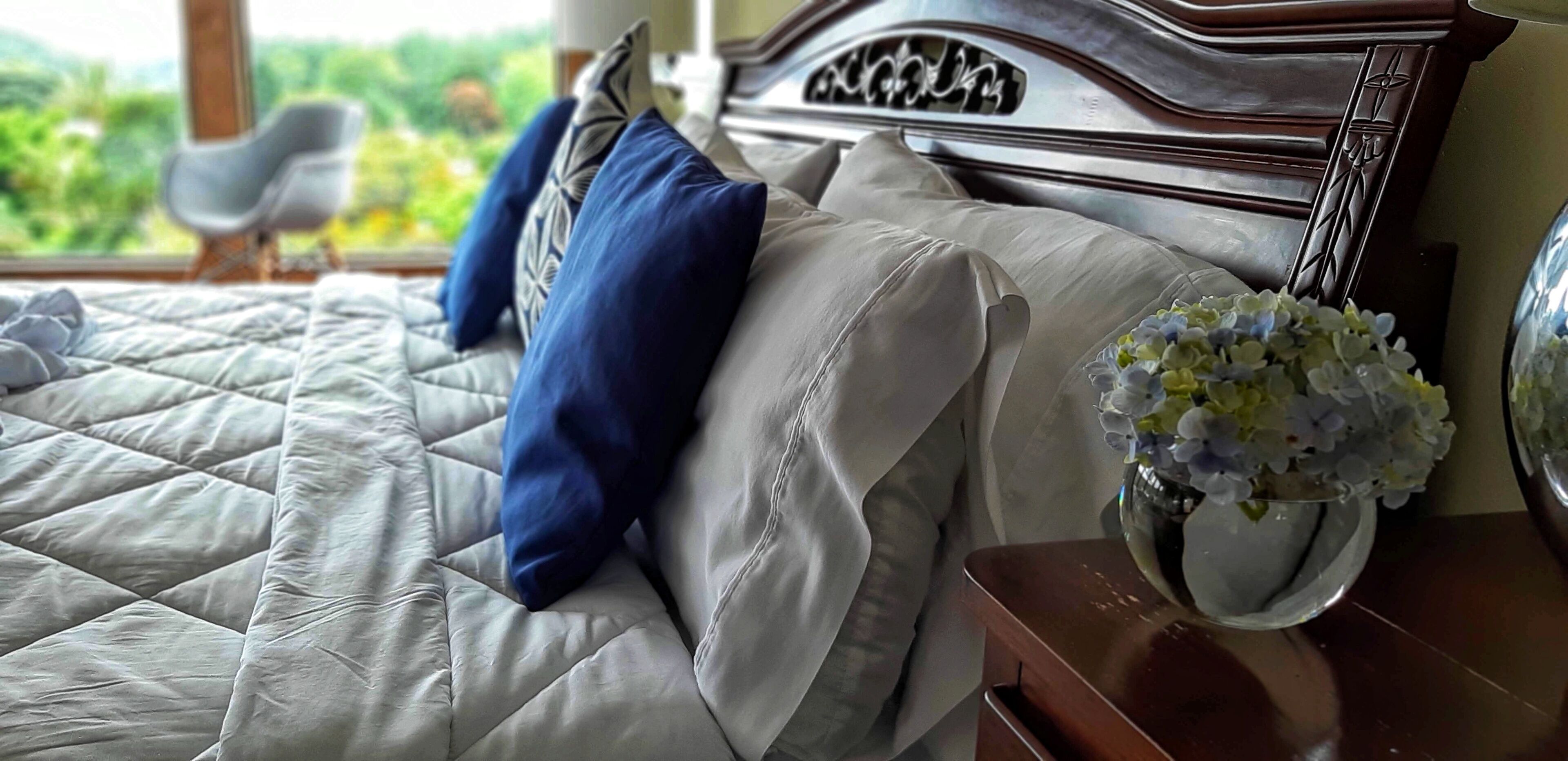 A neatly made bed with a quilted cover, a wooden headboard, and a vase with flowers on a bedside table.