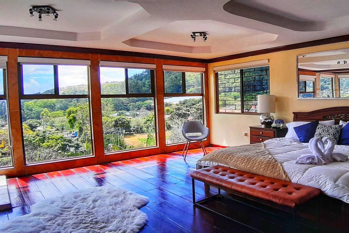 A spacious bedroom with a large bed, a bench, and a view of a lush landscape through large windows.