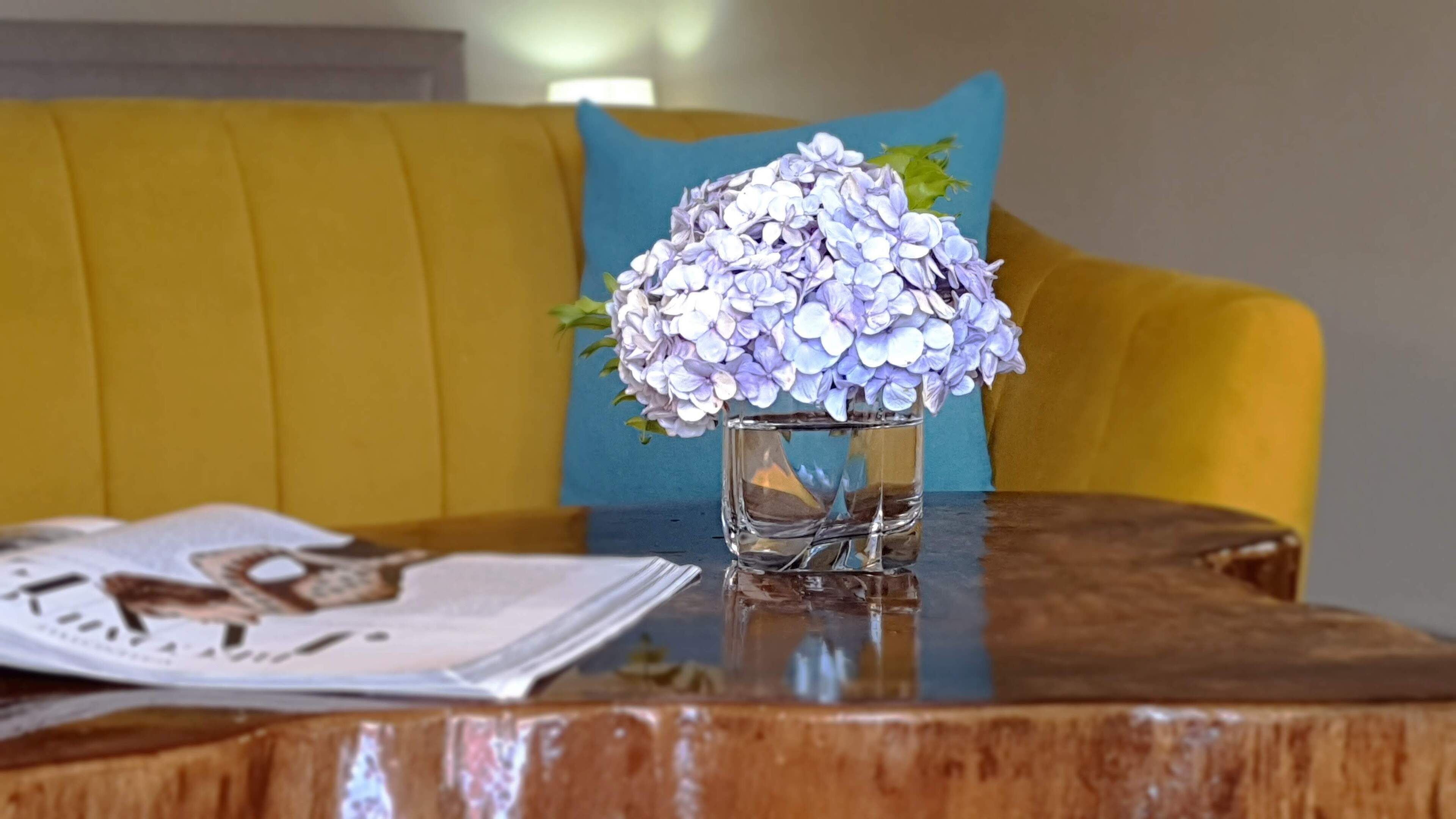 A wooden table with a vase of purple hydrangeas, a magazine, and a yellow couch with a blue pillow in the background.