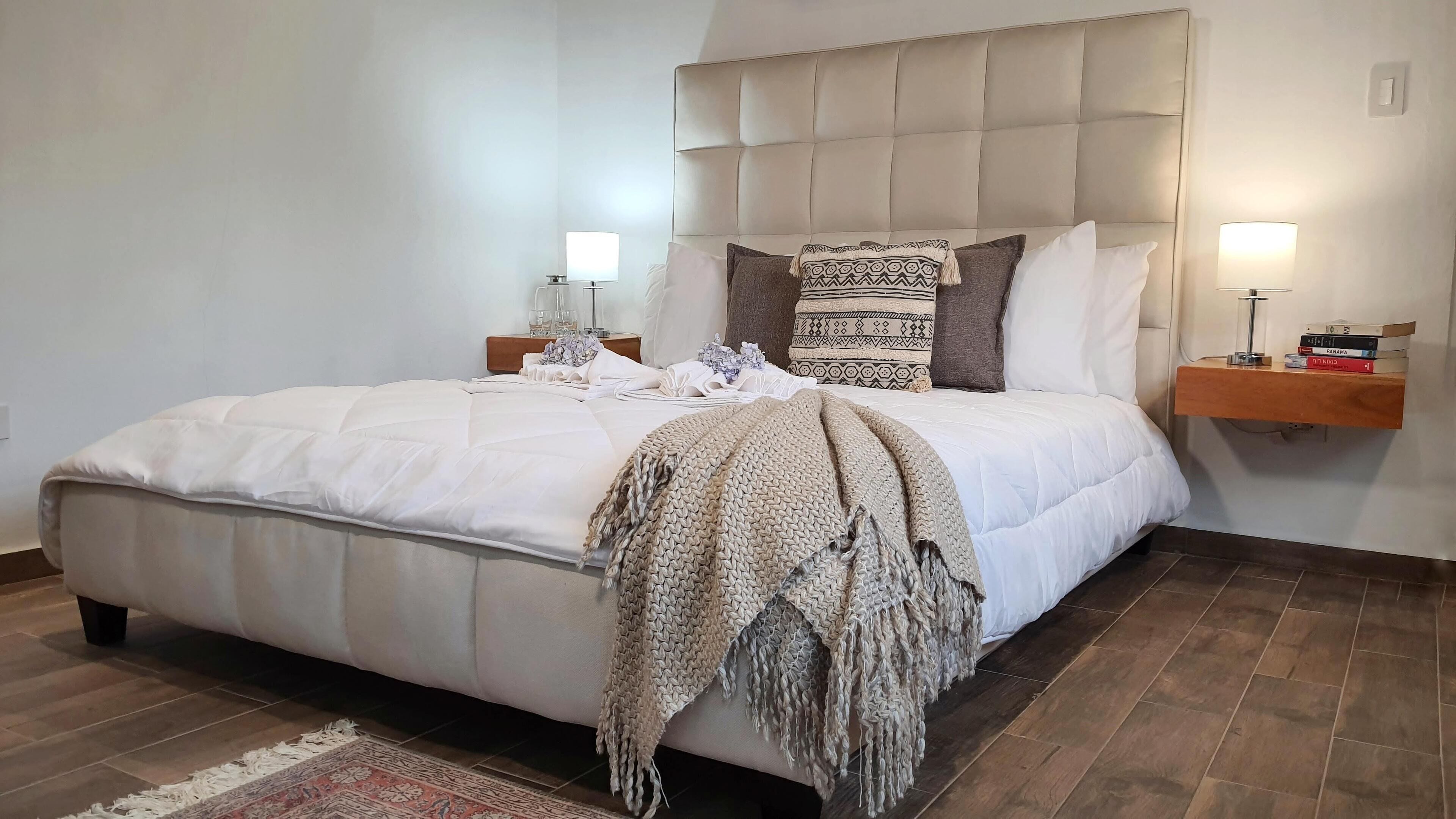 A neatly made bed with a white duvet, a knitted throw, and a patterned pillow. There are two bedside tables with lamps and a small shelf with books. The room has a wooden floor and a plain white wall.