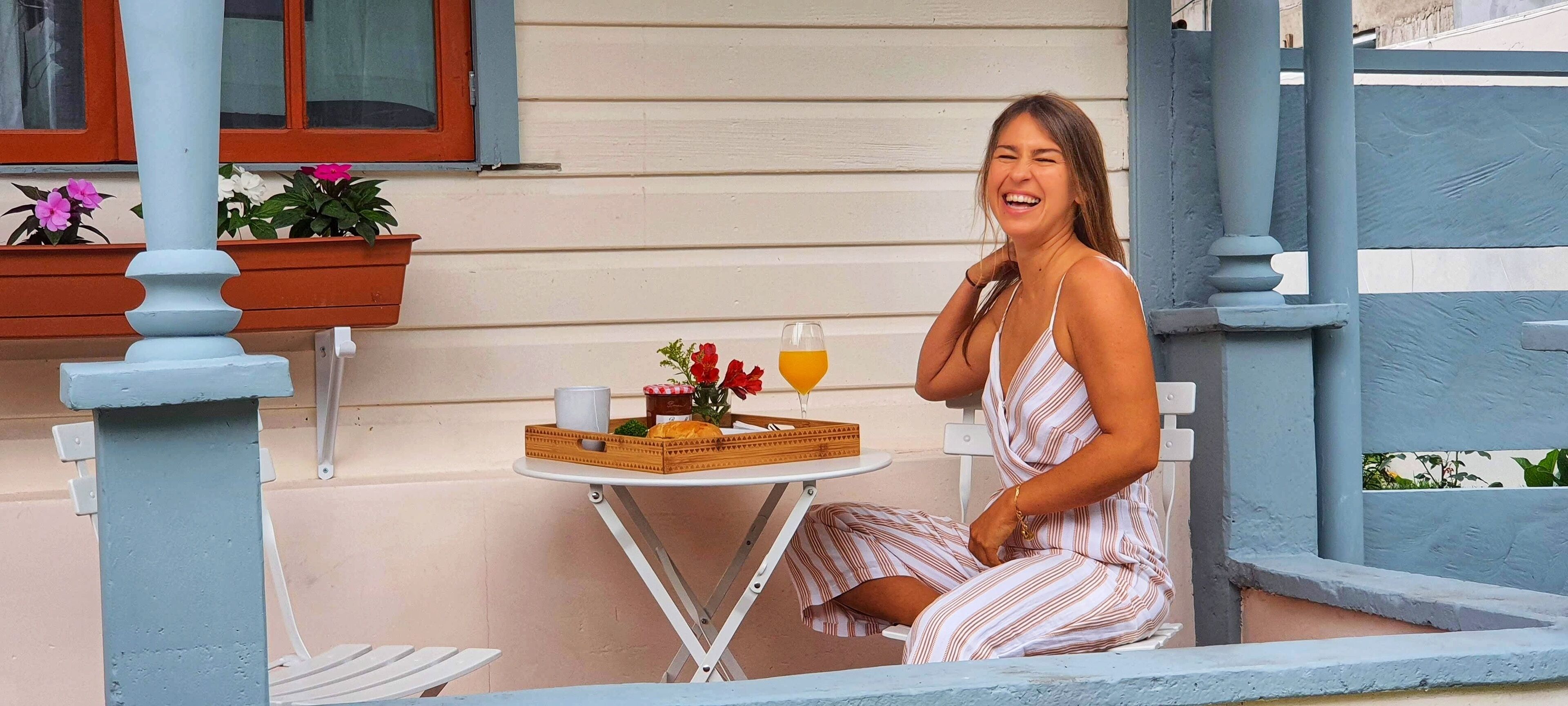Mulher sentada em pequena mesa com bandeja de comida e bebidas, janela com vista para a casa e um vaso com flores.