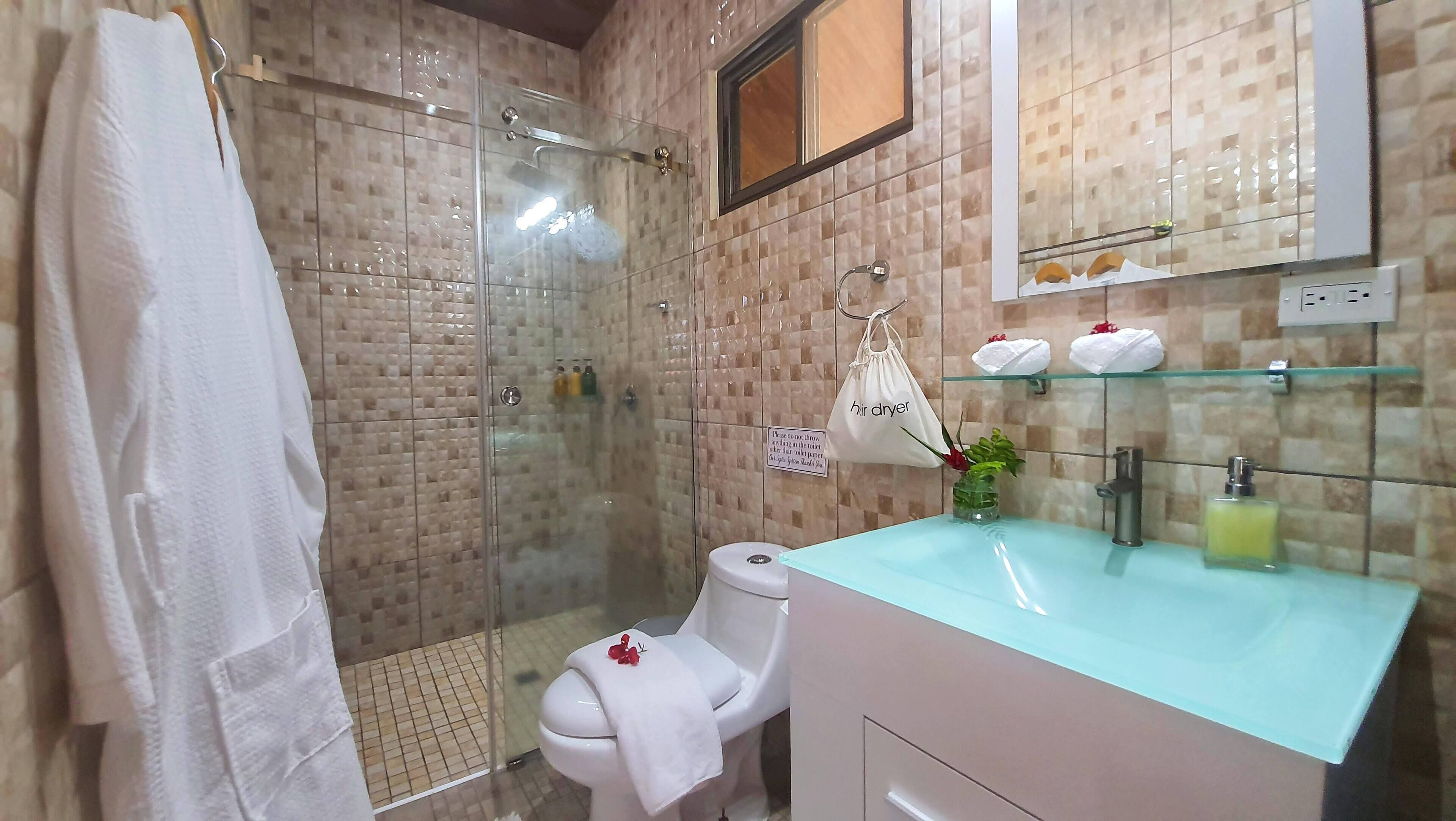 A bathroom with a glass shower enclosure, a toilet with a white towel, a sink with a glass countertop, and a mirror above the sink.